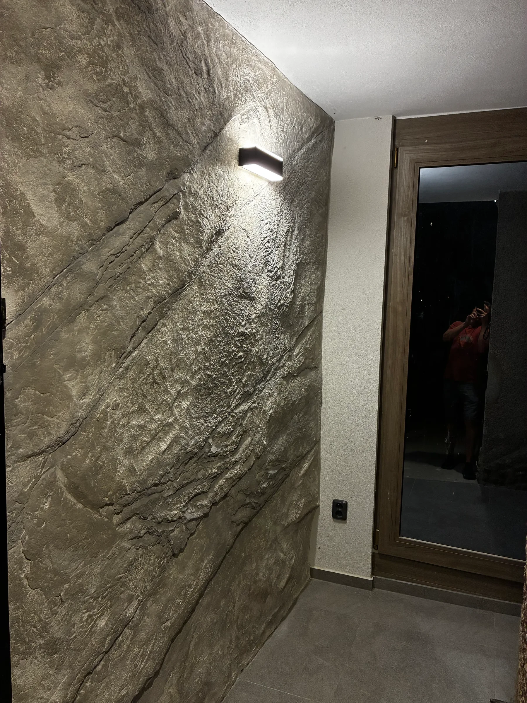 Stěna u&nbsp;vstupu večer, lampa svítí přímo ze skály dolů po reliéfu
